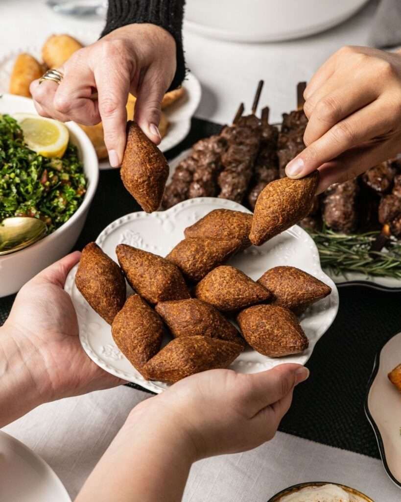 Mãos se servindo de kaftas e kibbehs em uma mesa compartilhada